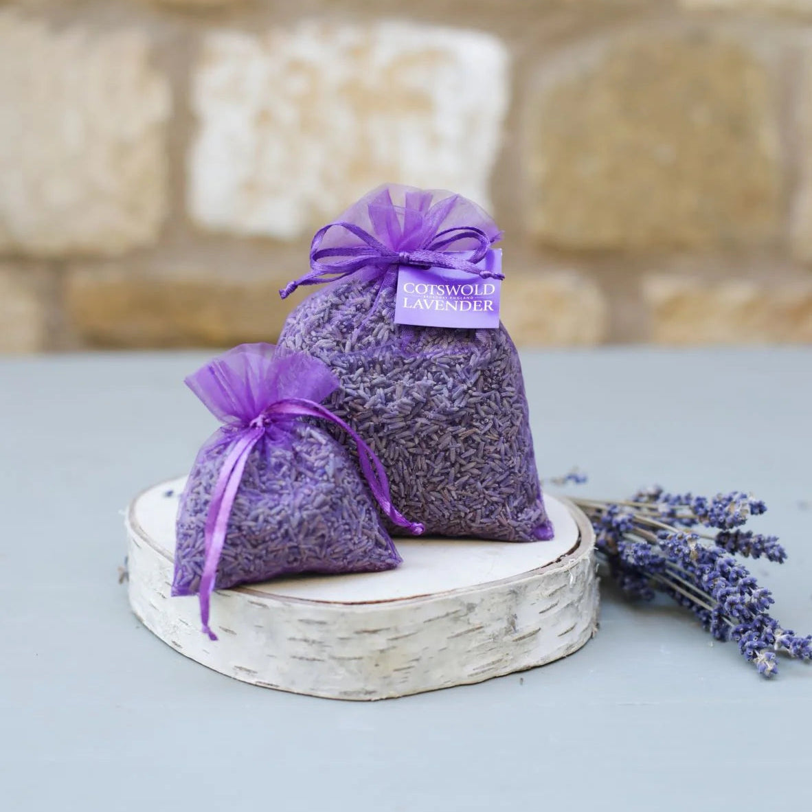Two lavender bags with purple ribbons on a stone surface with a blurred stone wall background.